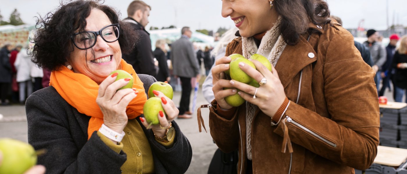 Två kvinnor med famnen full av äpplen skrattar och tittar på varandra. Foto.