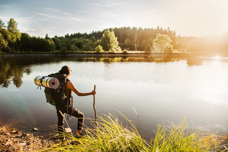 Ensam vandrare med ryggsäck och vandringsstav blickar ut över en sjö. Foto. 