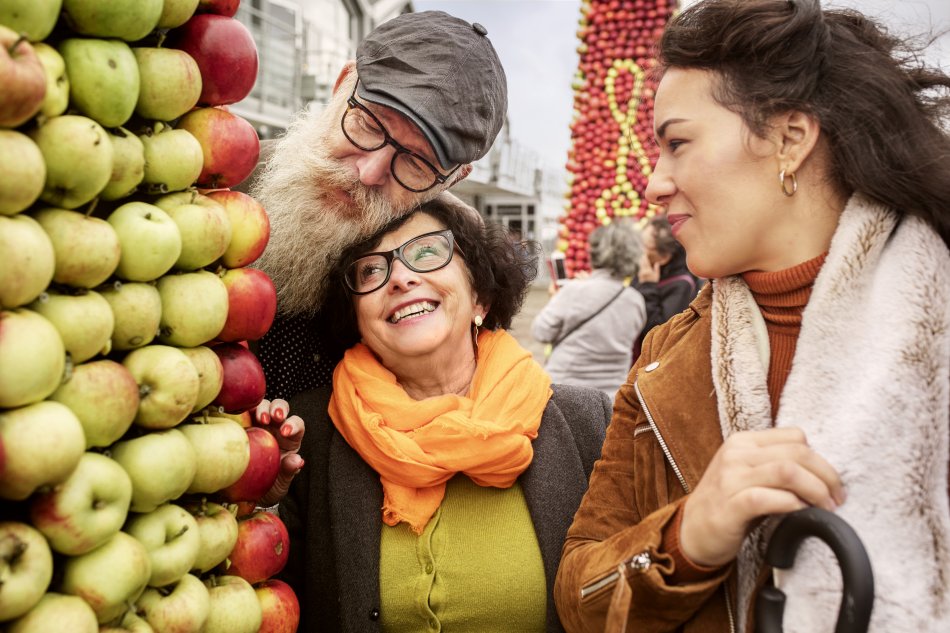 Tre glada personer står tätt intill varandra och tittar på en tavla gjord av äpplen. Foto.
