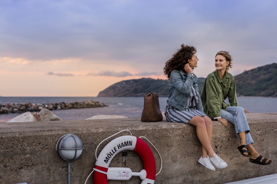 Två kvinnor sitter på en stenpir vid en hamn och pratar förtroligt med varandra. Foto.