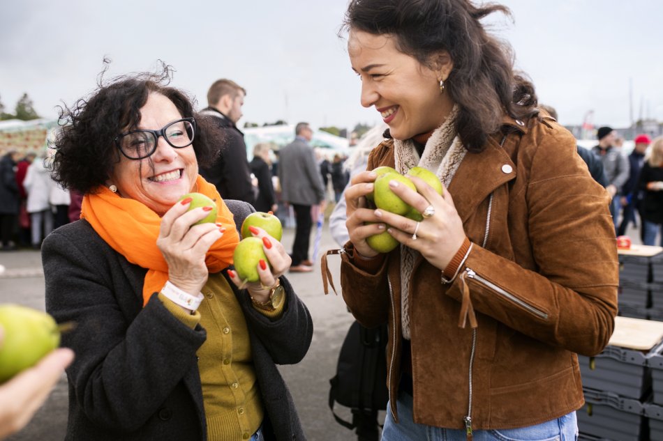 Två leende kvinnor håller äpplen hårt i sin famn. Foto.